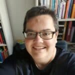 Porträtaufnahme einer jungen Frau mit kurzen Haaren, Brille und offenem Lächeln vor einem Bücherregal in legerer Kleidung.