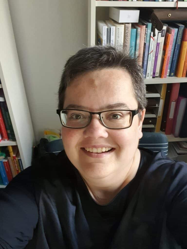 Porträtaufnahme einer jungen Frau mit kurzen Haaren, Brille und offenem Lächeln vor einem Bücherregal in legerer Kleidung.