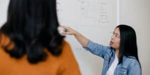 Female person pointing at white board and speaking.