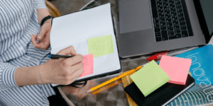 Top view of table with laptop and post-its, person writing into notebook